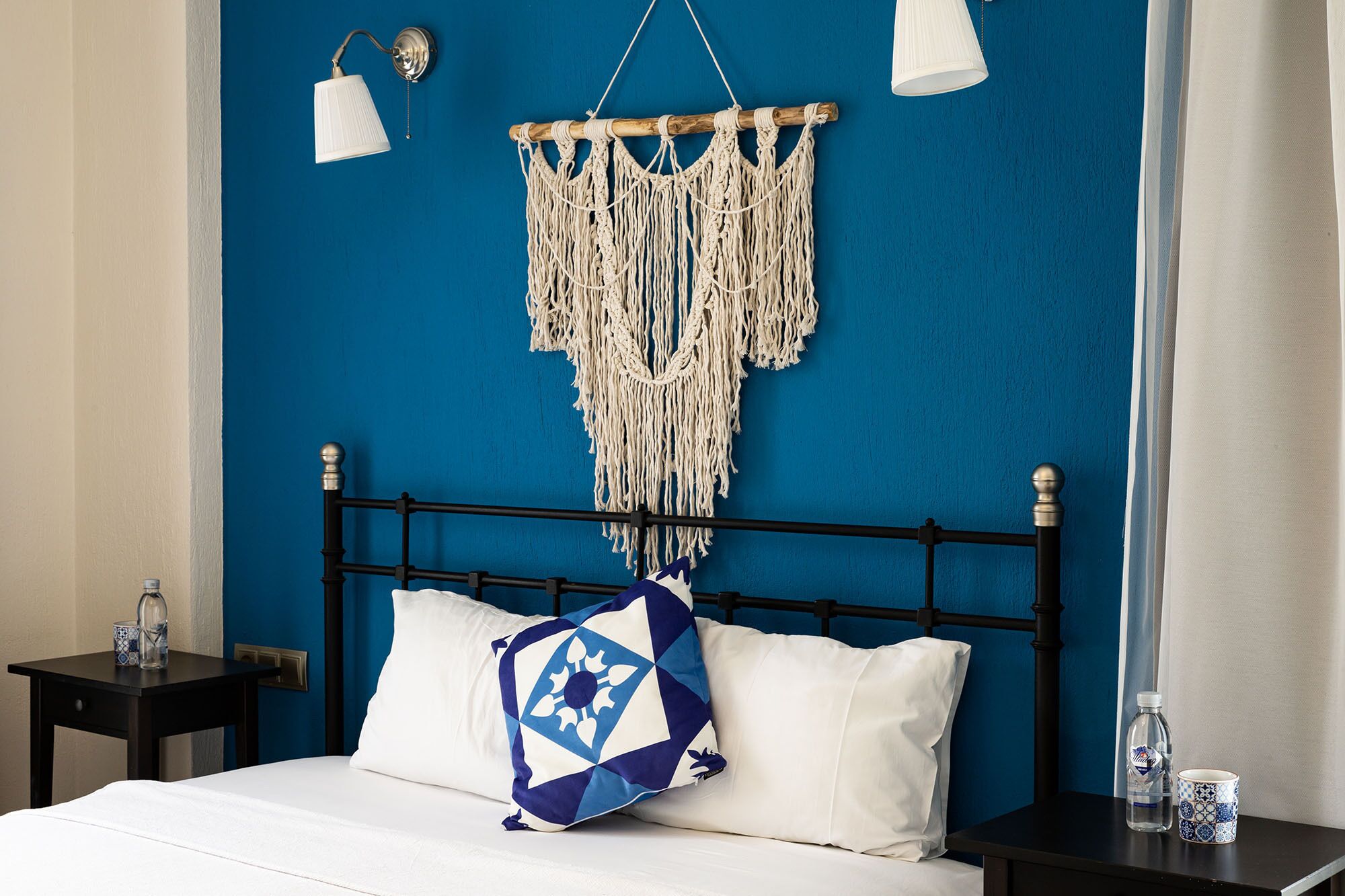Una cama con estructura metálica negra, un colgador de macramé y un cojín con estampado azul y blanco.