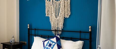 Una cama con estructura metálica negra, un colgador de macramé y un cojín con estampado azul y blanco.