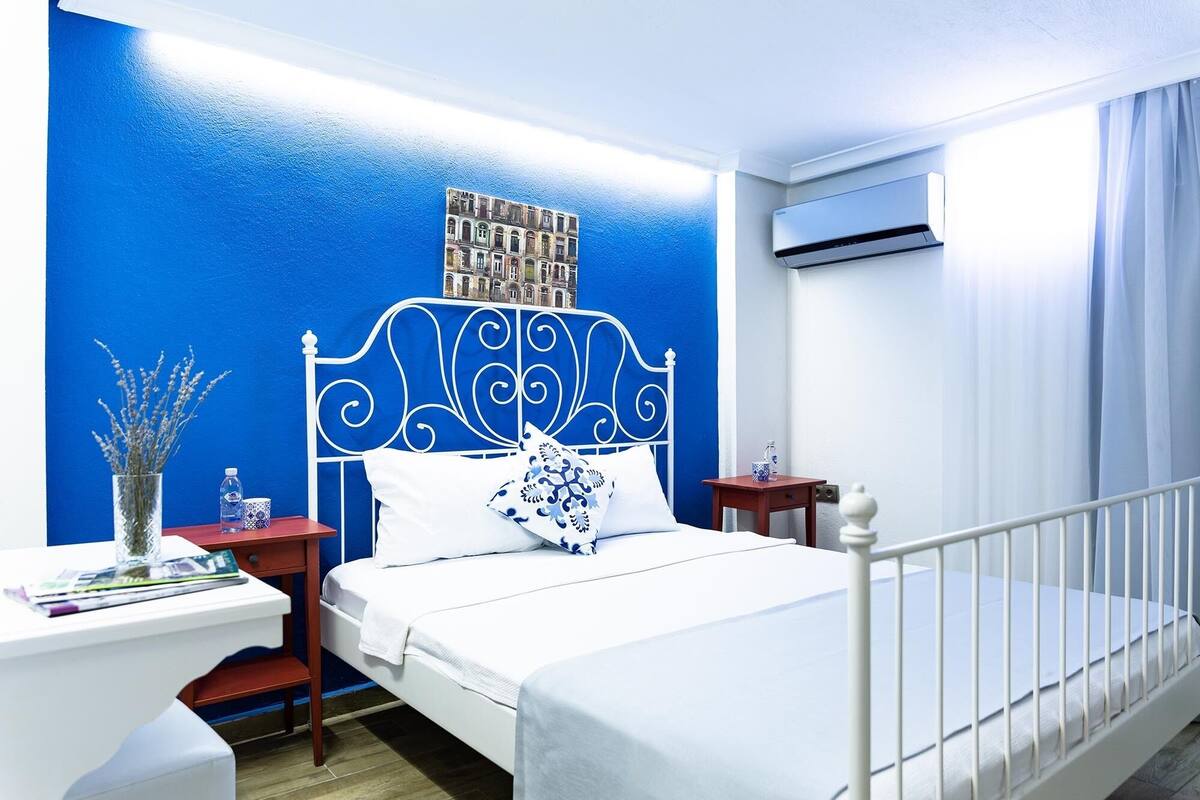 A bedroom with a white bed, a blue accent wall, a decorative headboard, a nightstand with a vase of flowers, and an air conditioning unit.
