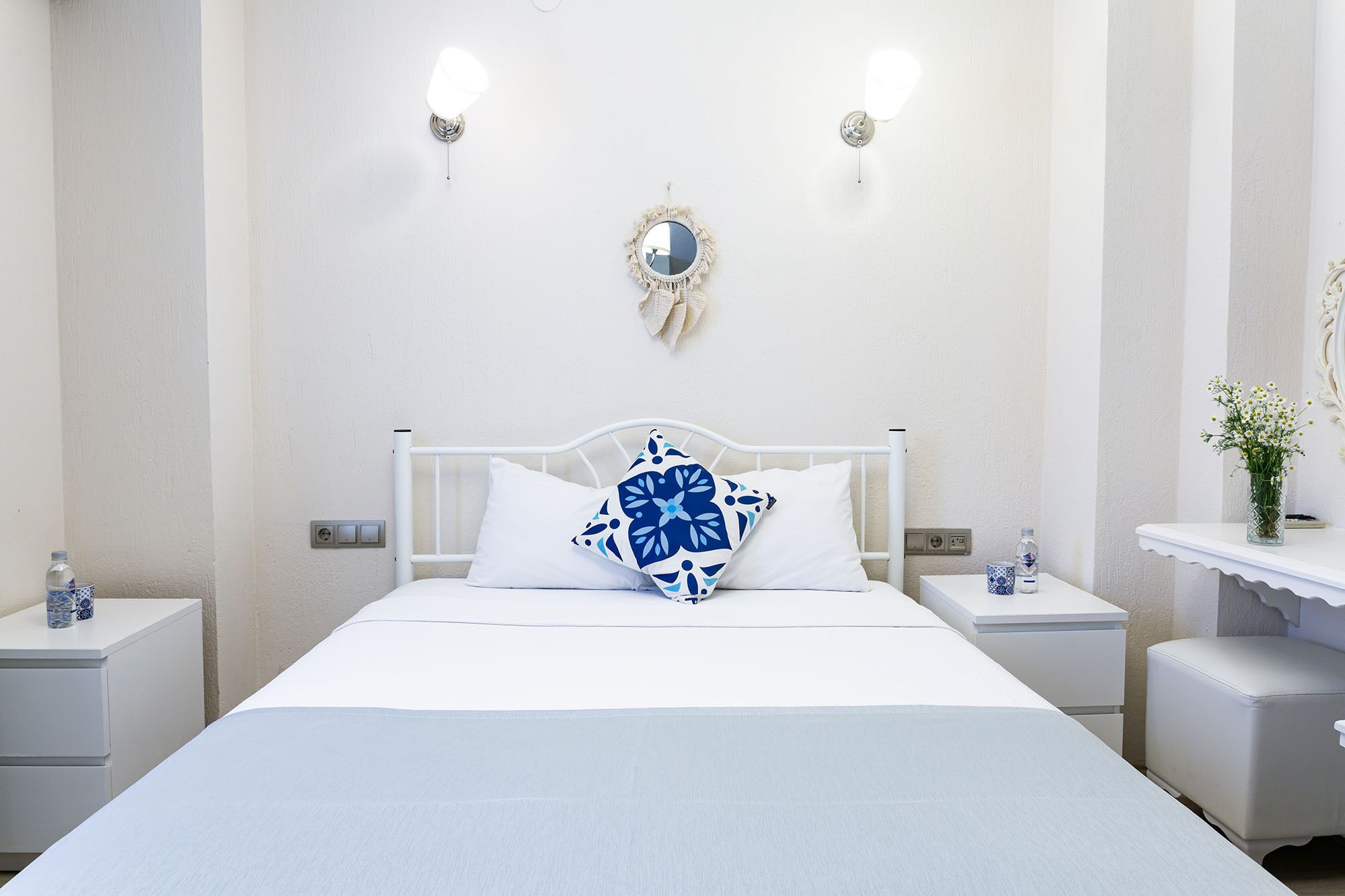 Een hotelkamer met een wit bed, twee nachtkastjes, een wandspiegel en een klein nachtkastje met een bloemenvaas.