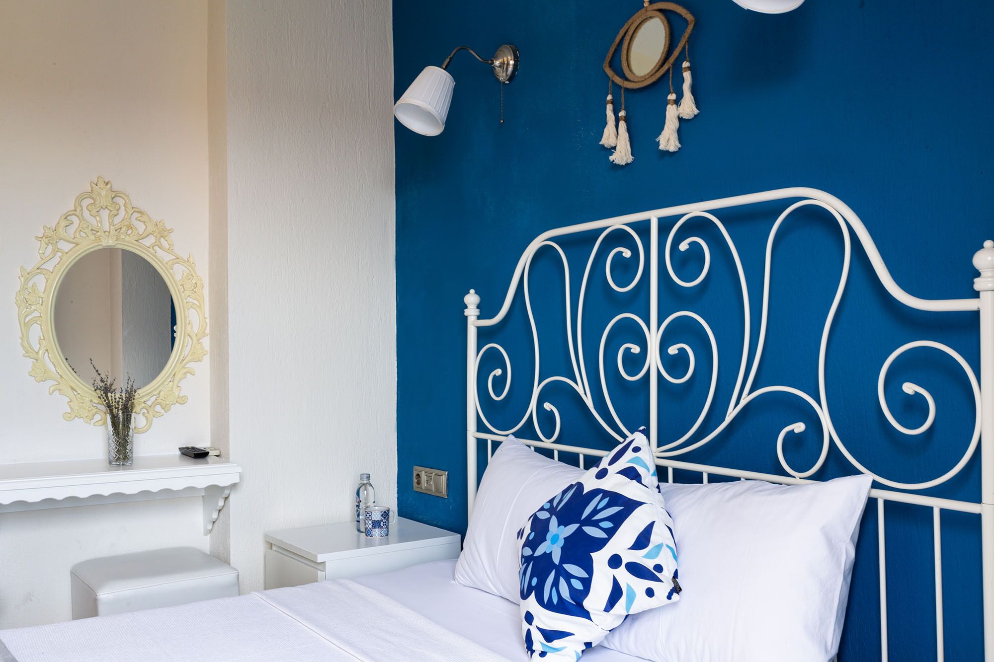 A bedroom with a white metal bed frame, a white bedside table, a white chair, a blue wall, and a decorative mirror.