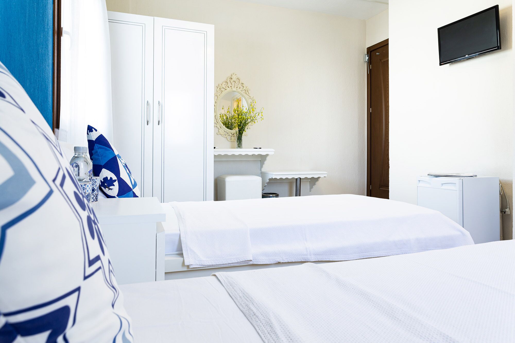 A hotel room with two beds, a white wardrobe, a small desk with a mirror, and a television mounted on the wall.