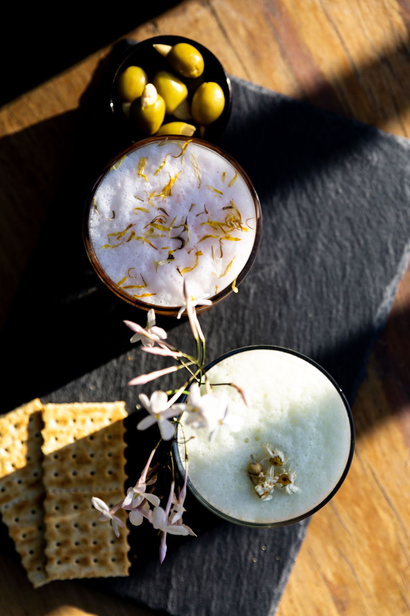 Three glasses of a white beverage with garnishes, accompanied by crackers and a bowl of yellow olives on a slate surface.