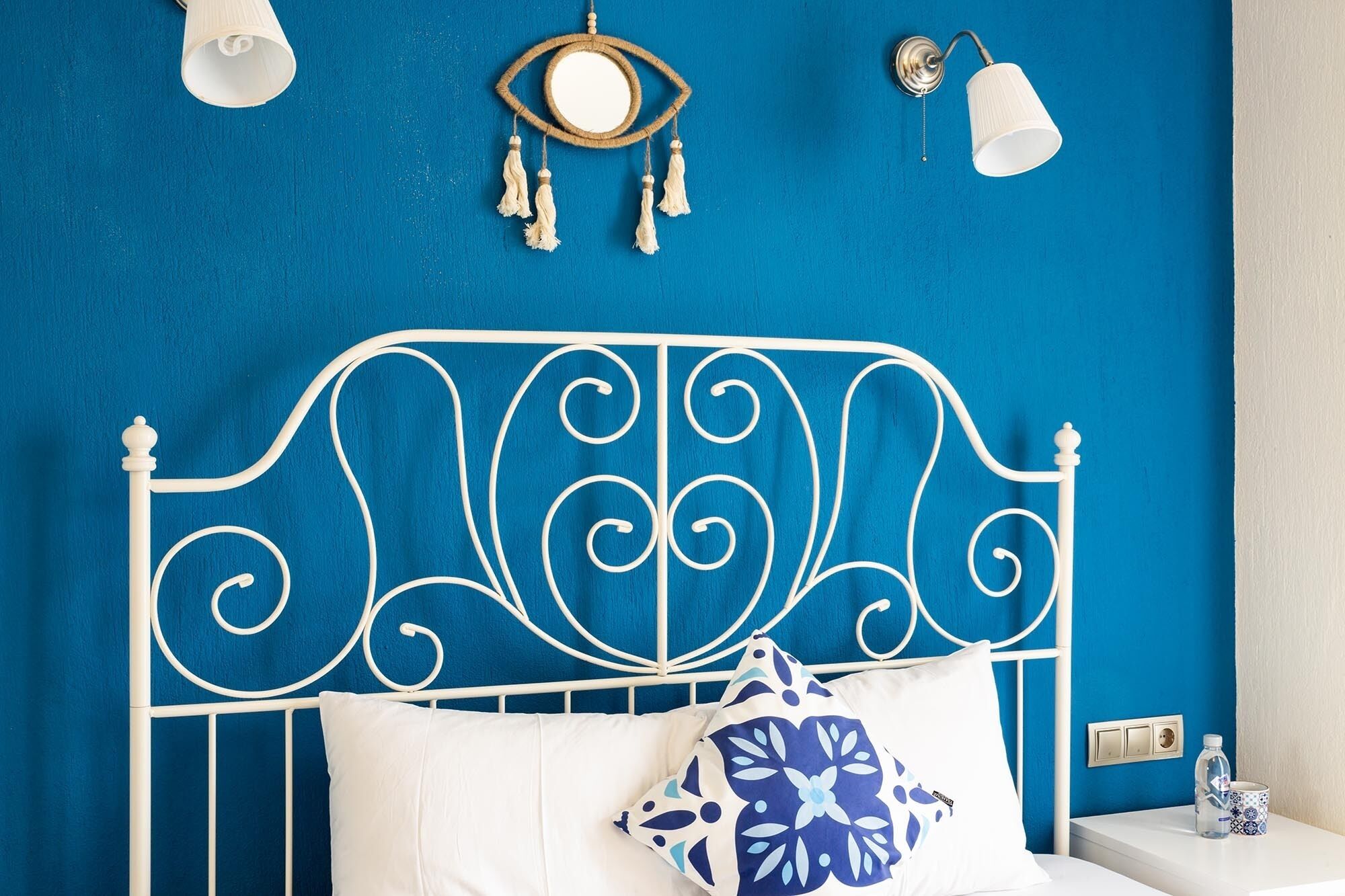 Una cama con cabecera metálica blanca, un almohadón blanco y un almohadón con estampado azul y blanco. Hay dos lámparas fijadas a la pared y un atrapasueños en la pared azul.