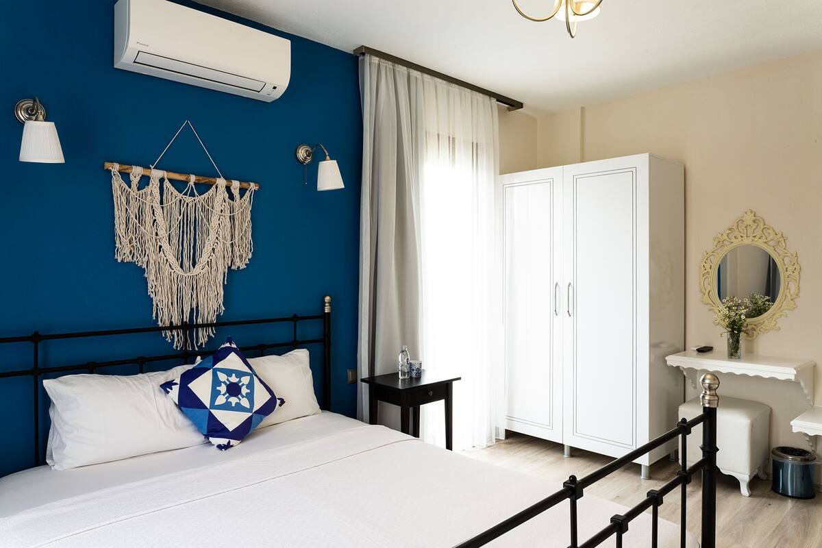 A bedroom with a bed, a wardrobe, a vanity with a mirror, and a macrame wall hanging.