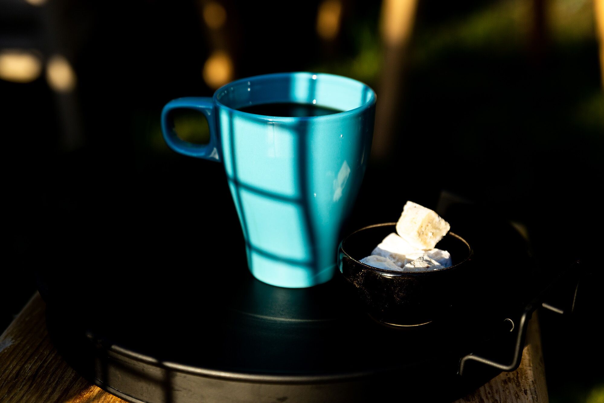 Une tasse bleue remplie d’un liquide sombre, probablement du café, est posée sur un plateau noir. À côté, un petit bol contient une substance blanche, vraisemblablement du sucre ou du sel.