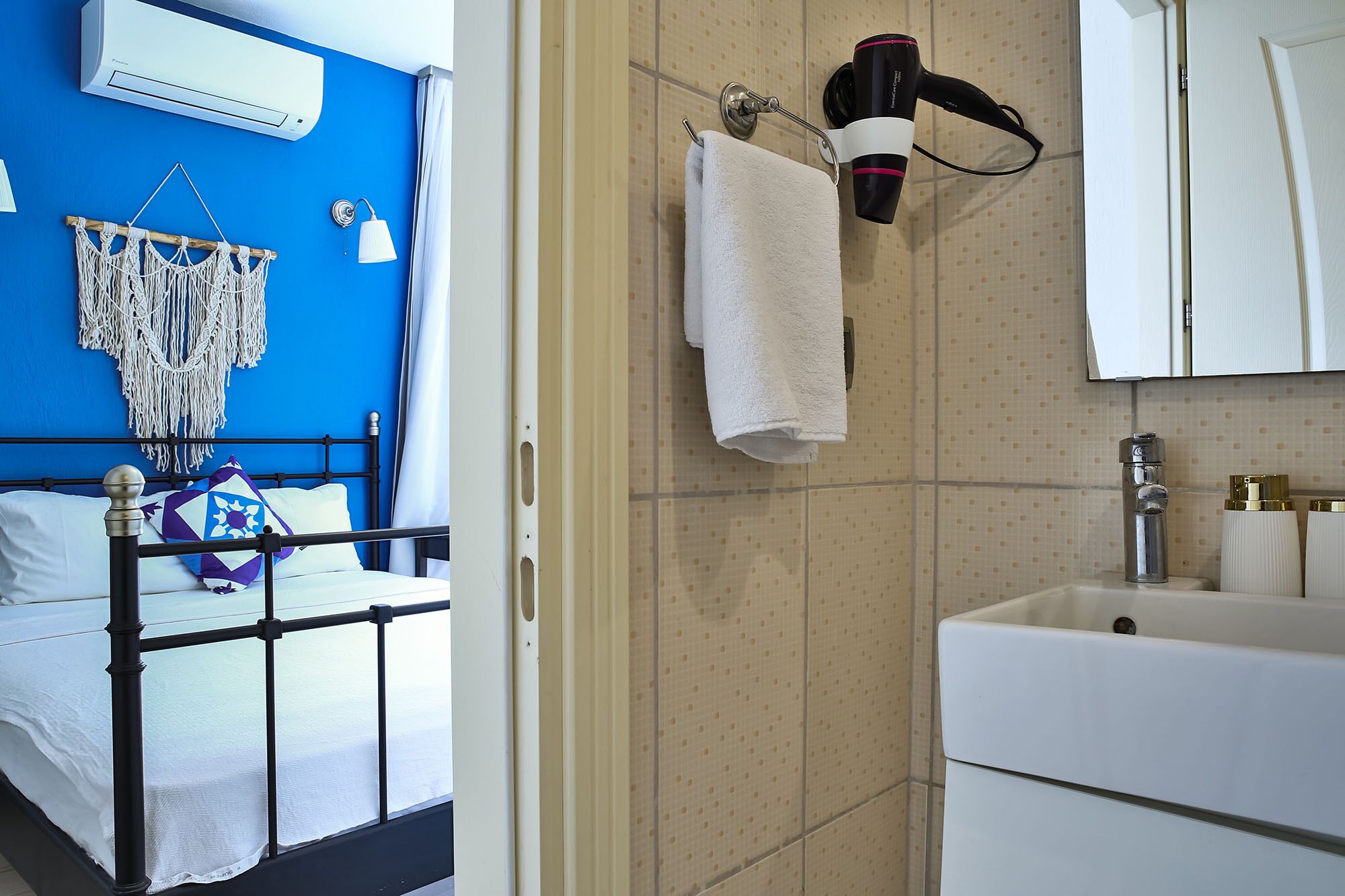 A room with a bed, a macrame wall hanging, a towel on a rack, and a bathroom with a sink and mirror.