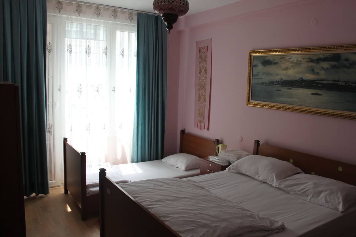 A bedroom with two beds, a window with curtains, a painting on the wall, and a nightstand.