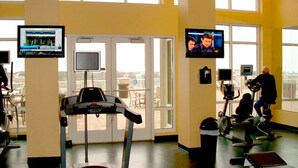 Ein Fitnessstudio mit Laufbändern, Ergometer und Kraftgeräten. Große Fenster und ein an der Wand befestigter Fernseher sind ebenfalls vorhanden.
