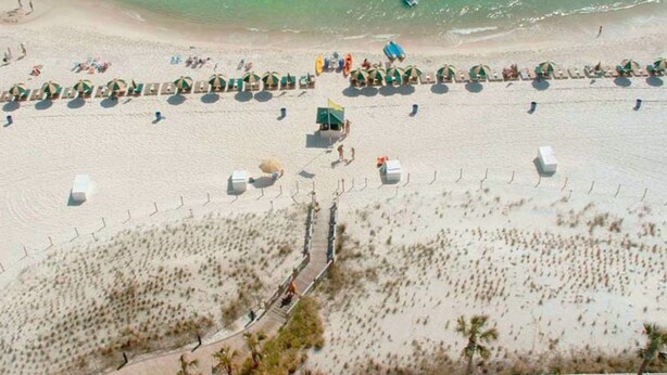 Luftaufnahme eines Strandes mit Reihen von Strandsonnenschirmen und Liegestühlen, einem Rettungsturm und einer sandigen Küstenlinie mit spärlicher Vegetation.