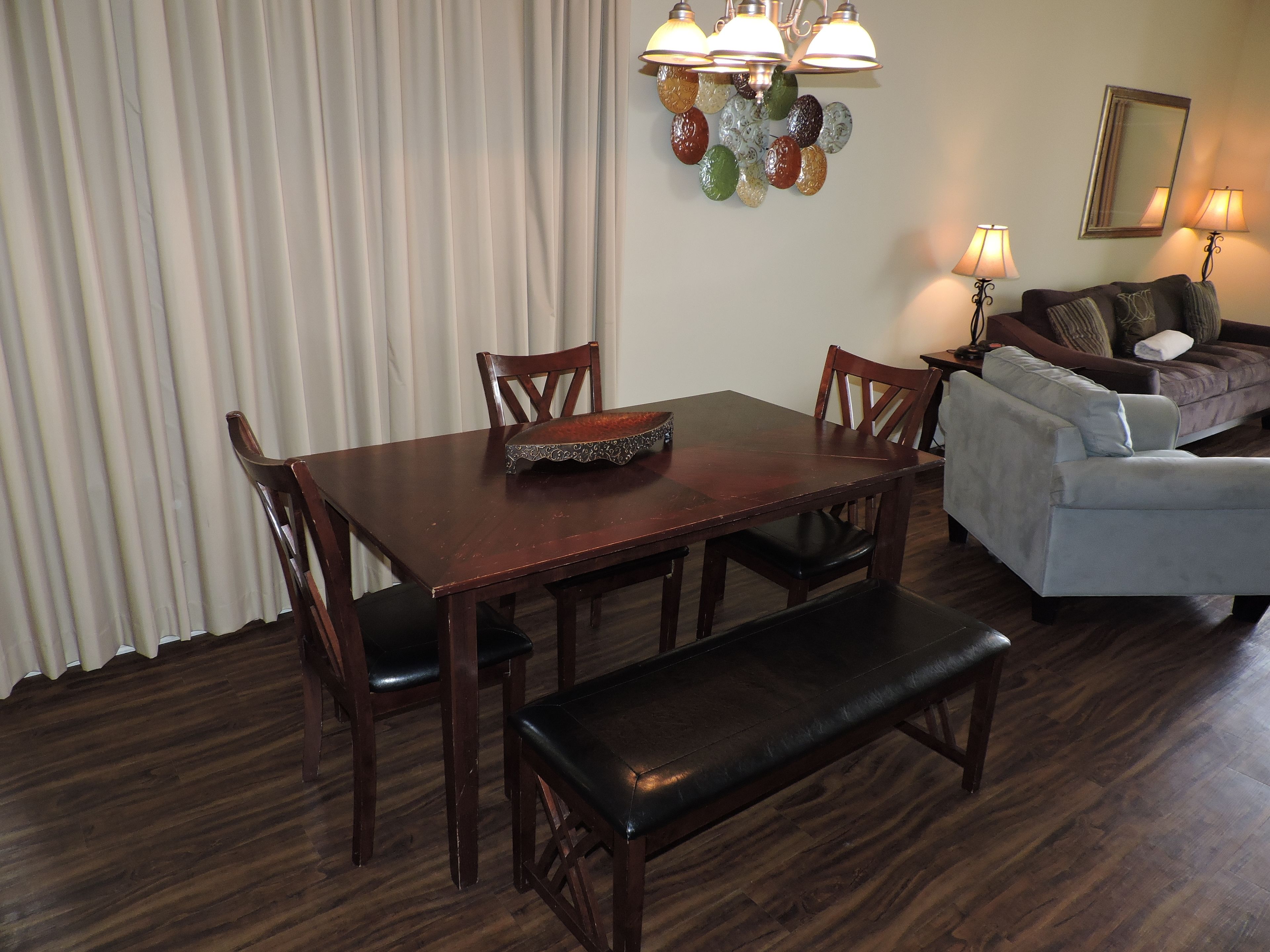 A dining area with a wooden table, chairs, and a bench. There is a sofa and a lamp in the background.