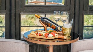 Una mesa con una botella, vasos y un plato de comida cerca de una ventana que da a vista de barcos.