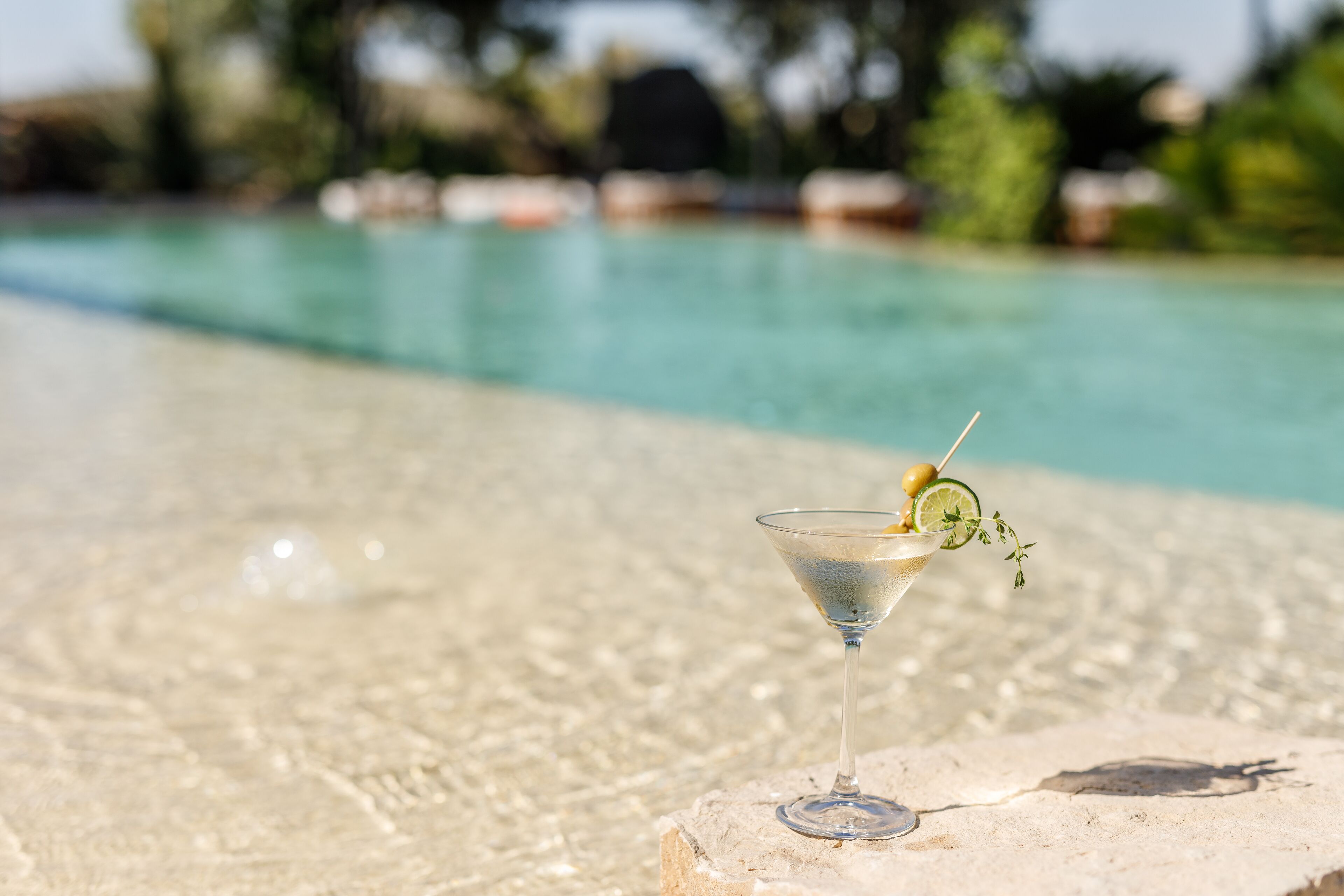 Ein klarer Martini in einem Glas, garniert mit Limettenspalte und Olive, auf einer Steinfläche neben einem Pool.
