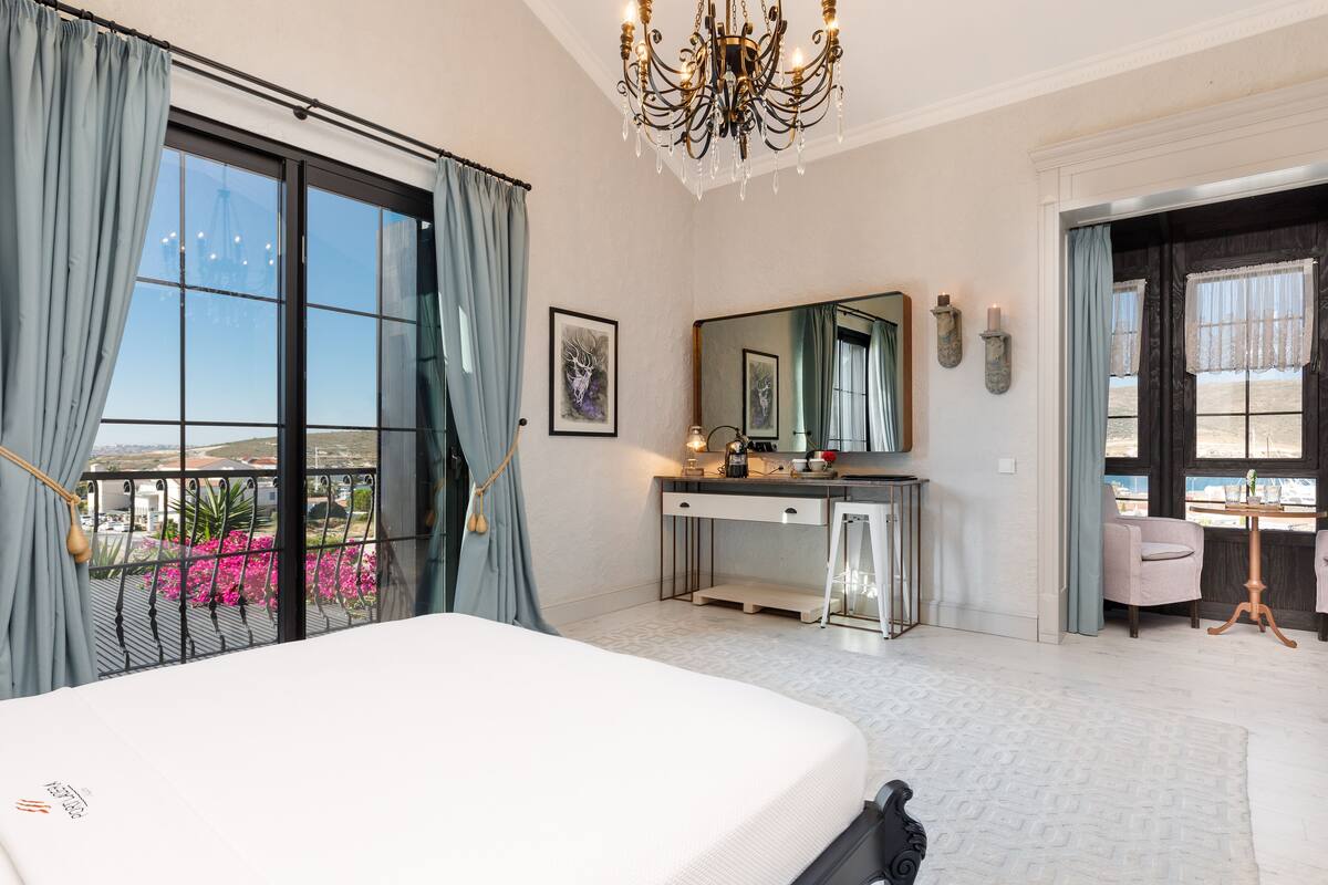 Un dormitorio con una cama grande, un tocador con espejo, un escritorio y una silla. Hay un balcón con vistas a la ciudad.