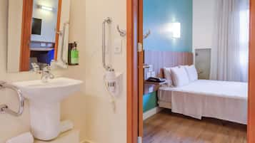 A hotel bathroom with a sink, mirror, and toilet. There is a bed in the adjacent room.