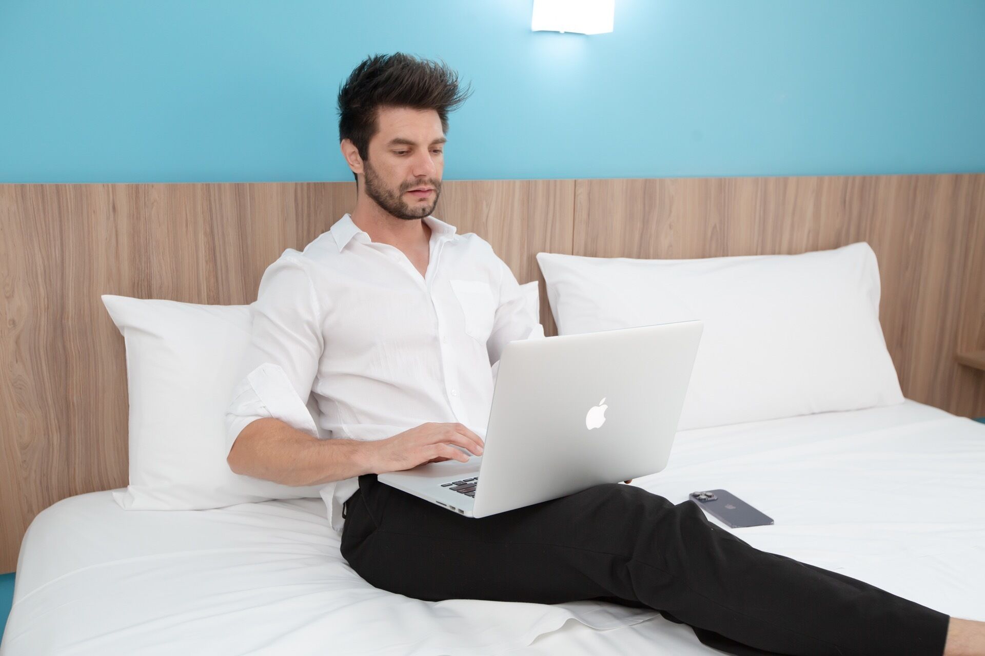 Una persona sentada en una cama con una computadora portátil, almohadas y un teléfono inteligente.