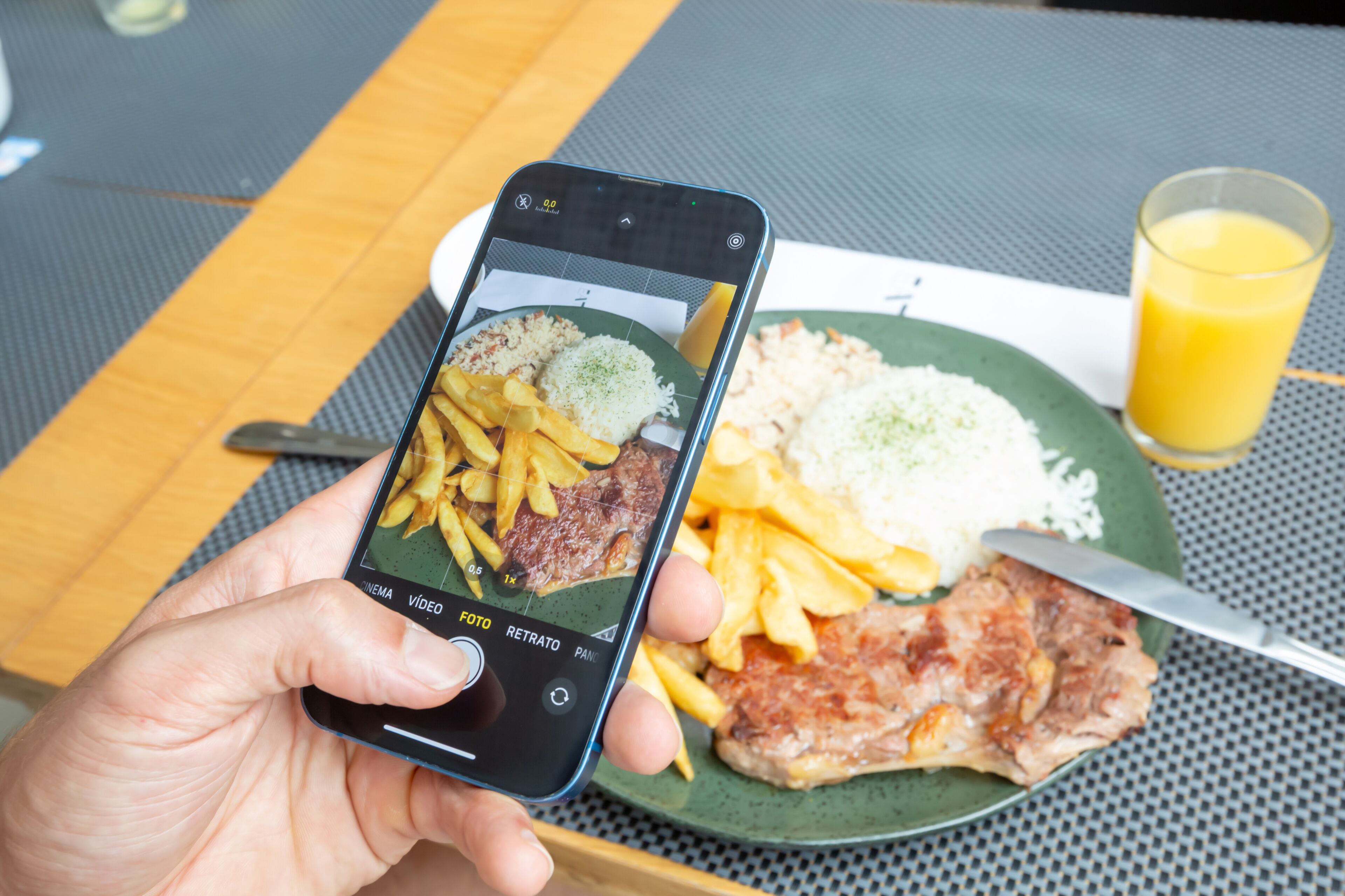 A person holding a smartphone with a photo of a meal on a green plate, including rice, fries, and a piece of meat, with a glass of orange juice on the table.