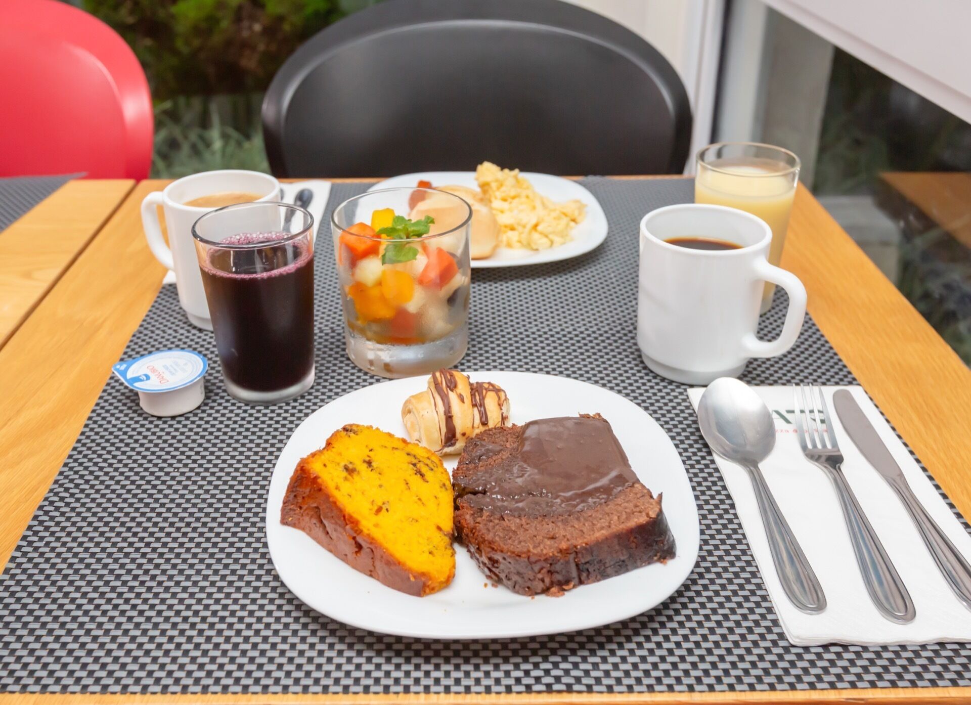 A table with a plate of food, a glass of dark liquid, a glass of yellow liquid, a cup of coffee, and a small container of yogurt.