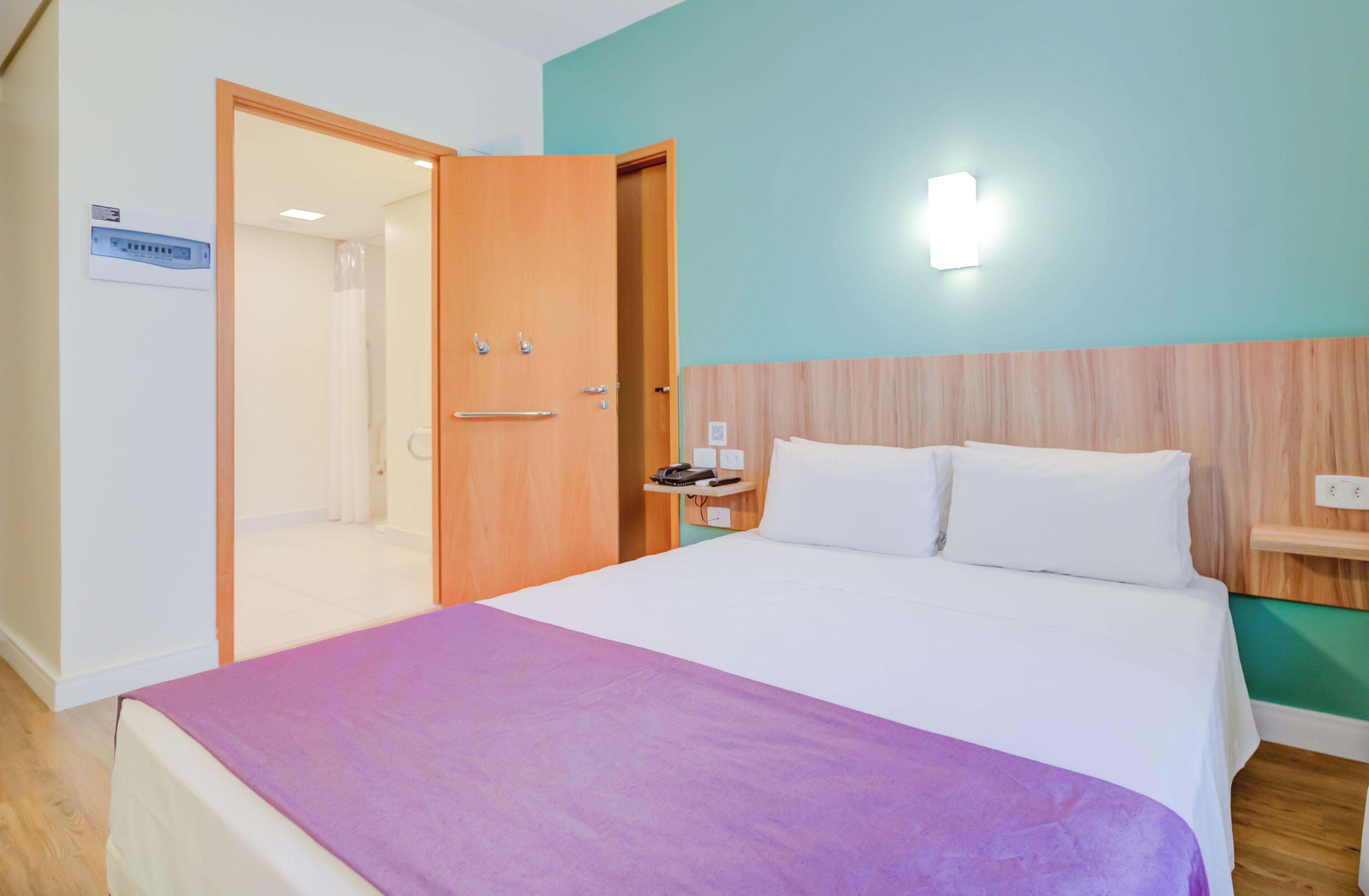A hotel room with a bed, a nightstand, a wooden headboard, a wall-mounted light, and a door leading to a bathroom.
