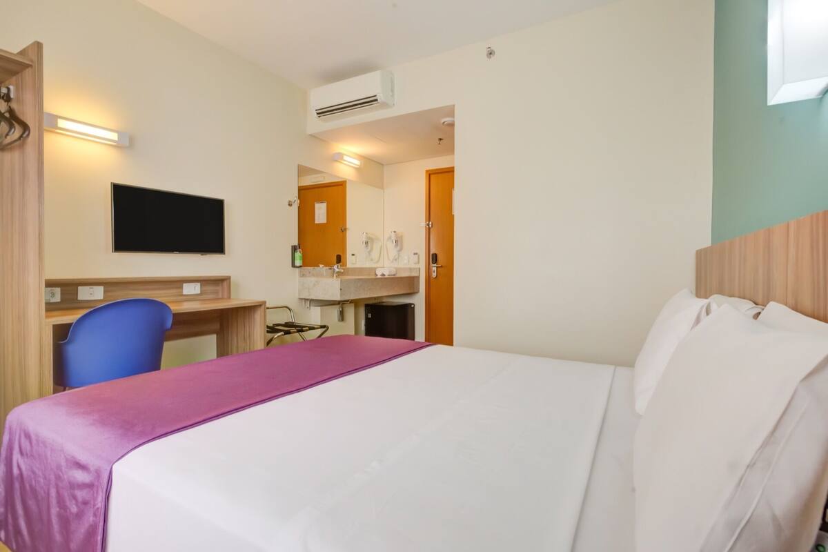 Une chambre d’hôtel comprenant un lit, un bureau avec une chaise, une télévision, une salle de bain avec un miroir et un appareil de climatisation.
