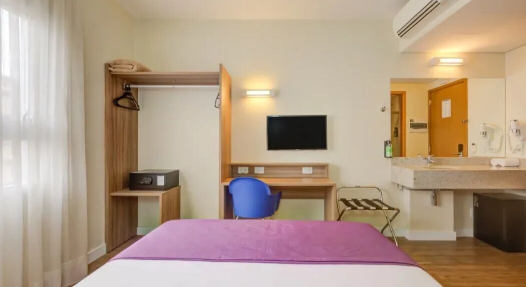 Una habitación de hotel con cama, escritorio, silla, televisión, espejo y baño.