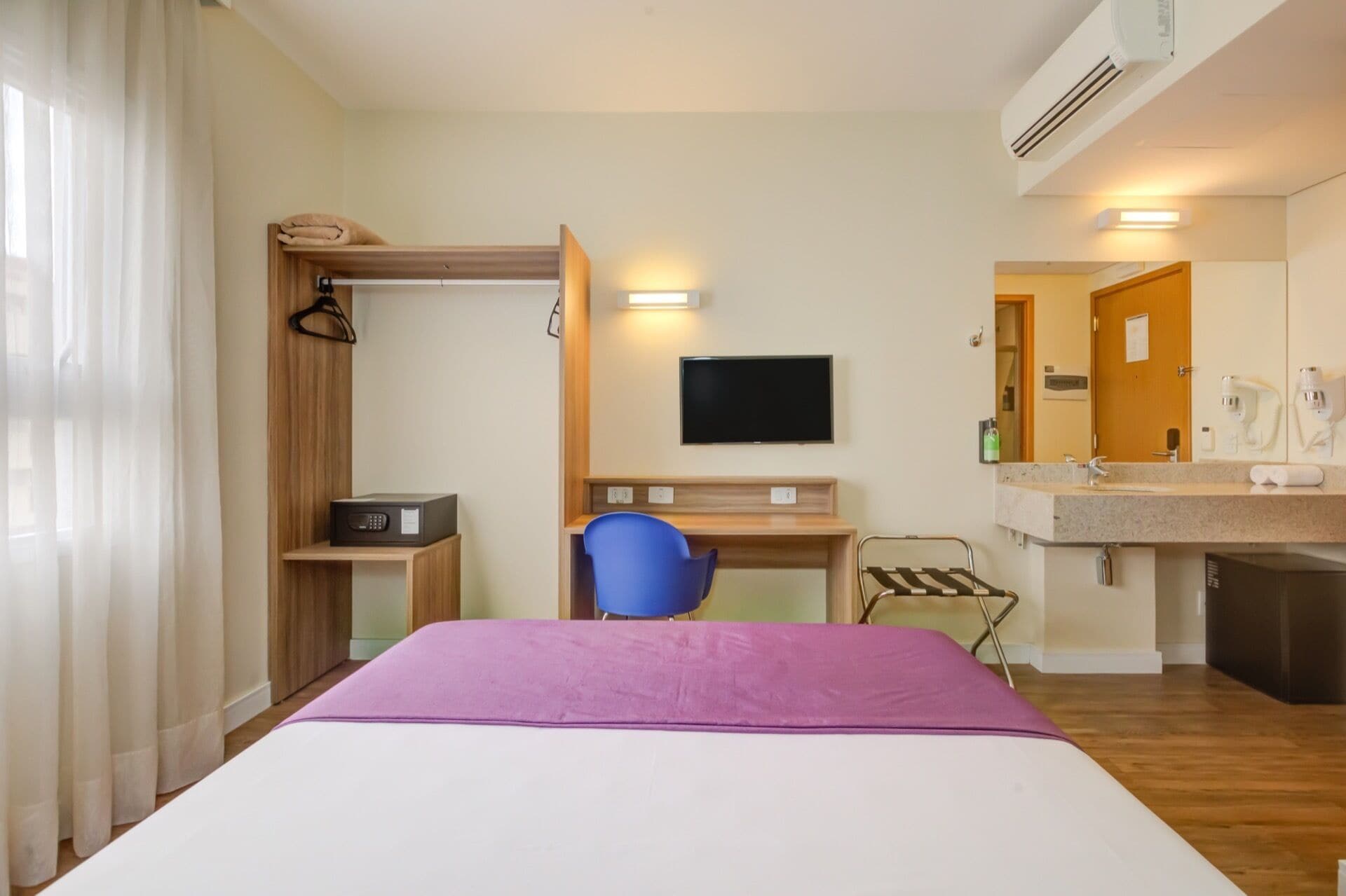 A hotel room with a bed, a desk with a chair, a TV, a mirror, and a bathroom with a sink and shower.