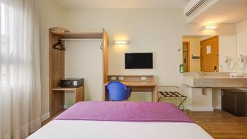 A hotel room with a bed, a desk with a chair, a TV, a mirror, and a bathroom with a sink and shower.