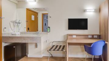 A hotel room with a wooden desk, a blue chair, a flat-screen TV, a small refrigerator, a sink with a mirror, and a towel rack.