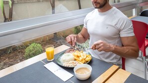 A man dining outdoors with a meal of rice, fries, and a beverage, with a view of greenery and a building in the background. - Transamerica Fit Campinas (Campinas)