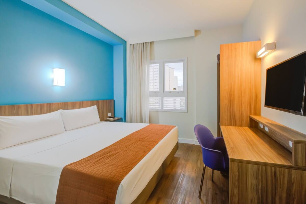 Une chambre d’hôtel avec un lit, un bureau, une chaise, une télévision et une fenêtre avec des rideaux.