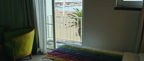 A hotel room with a bed, a green chair, a balcony with a view of the beach and marina.