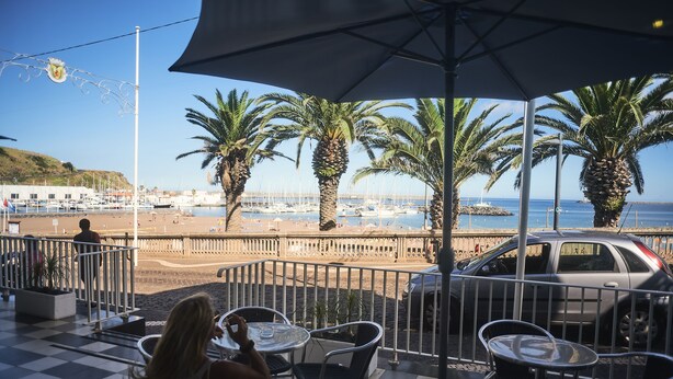 Terraza con mesas y sillas que da a una playa con palmeras y barcos.
