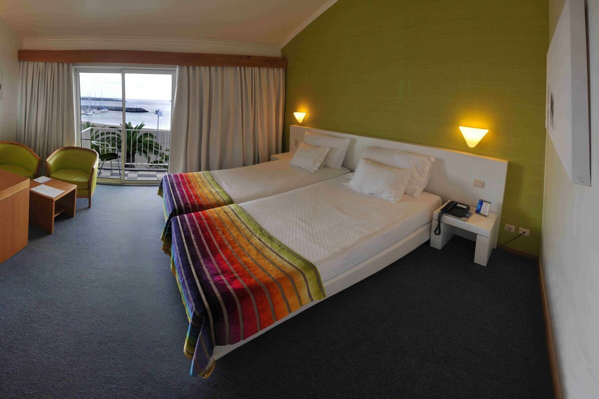 A hotel room with two beds, a desk, a chair, a nightstand with a phone, and a view of the sea through a large window.