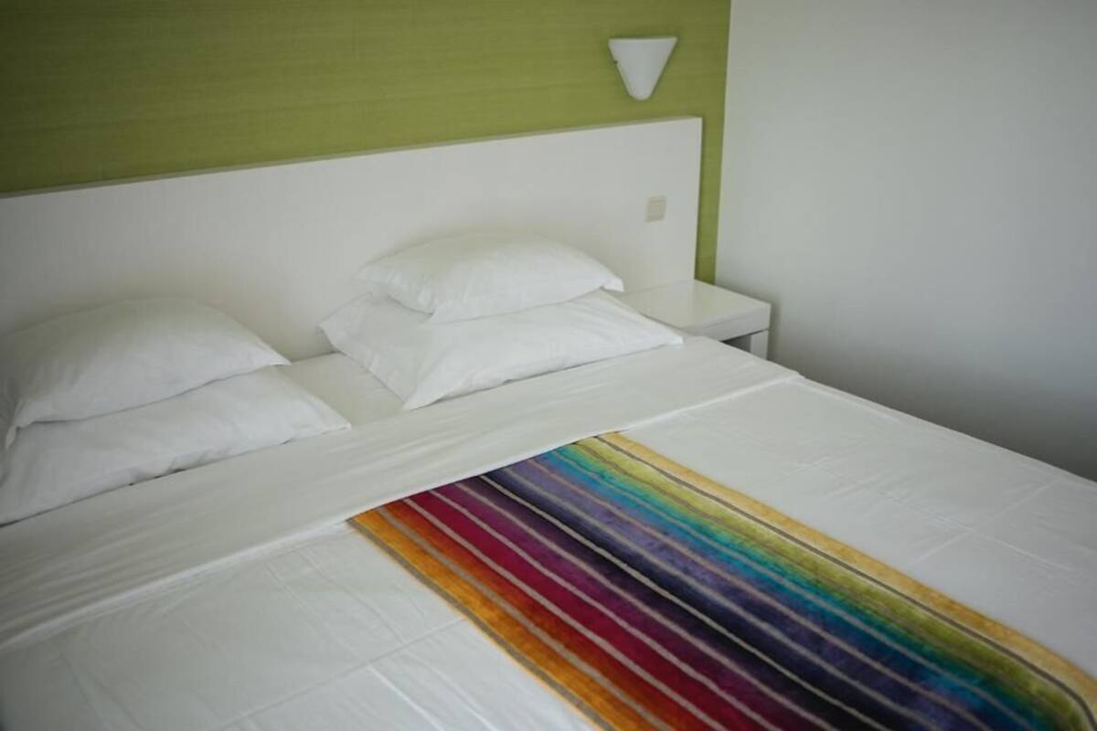 A bed with white bedding and a rainbow-striped blanket, a white nightstand, and a green wall with a white light fixture.