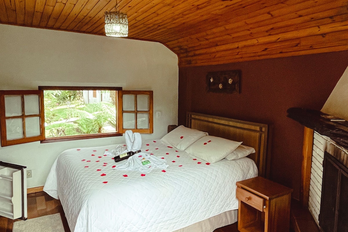 A bedroom with a wooden ceiling, a bed with rose petals, a nightstand, a fireplace, and a window with a view of greenery.