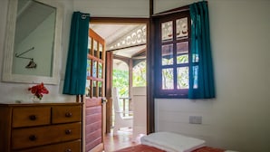 A room with a wooden dresser, a bed with a white blanket, a window with teal curtains, and a view of a garden through the open door. - Bogles Round House (Hillsborough)
