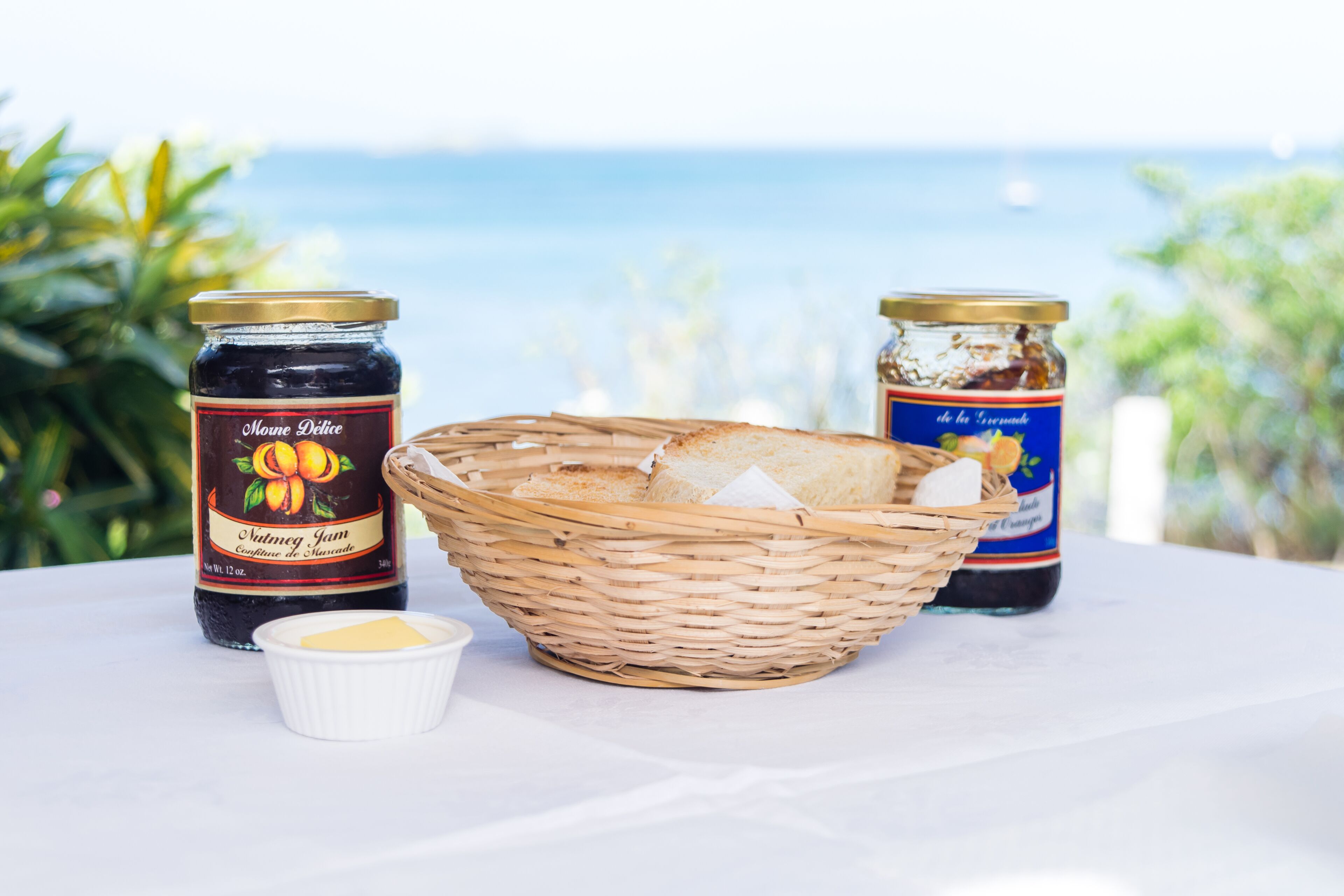 A table with two jars of jam, a basket with bread, and a small bowl of butter, set against a scenic view.