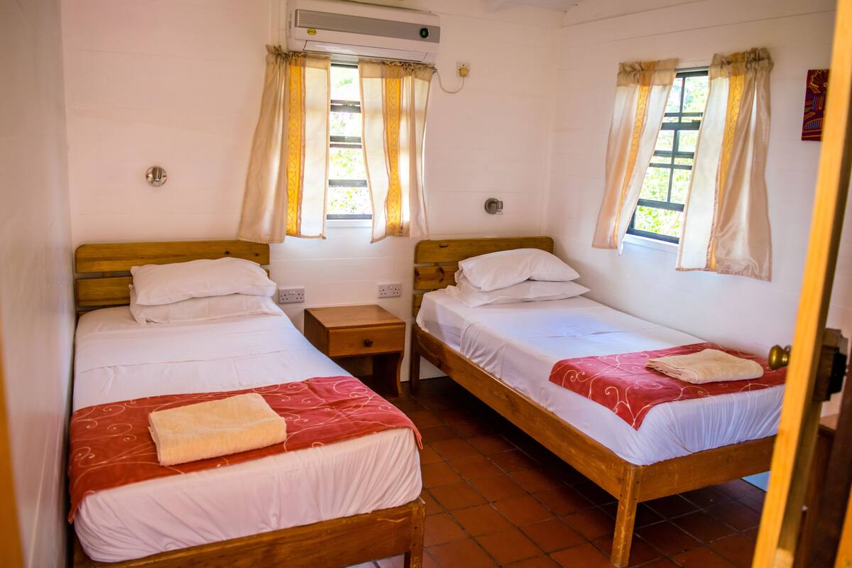 A room with two wooden beds, each with white bedding and a red patterned bedspread. There are two bedside tables, one on each bed, and a window with curtains.