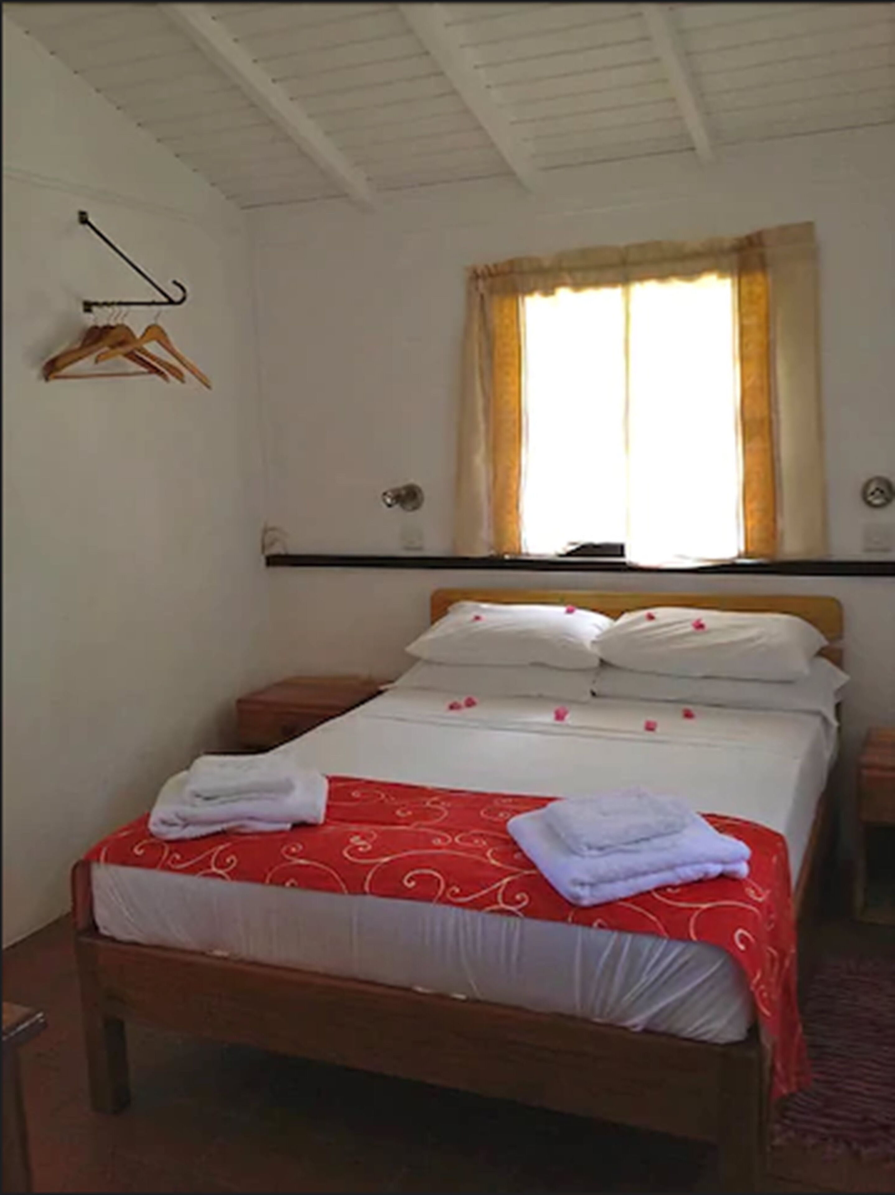 A neatly made bed with a red and white bedspread, two pillows, and two folded towels. A wooden nightstand is beside the bed.