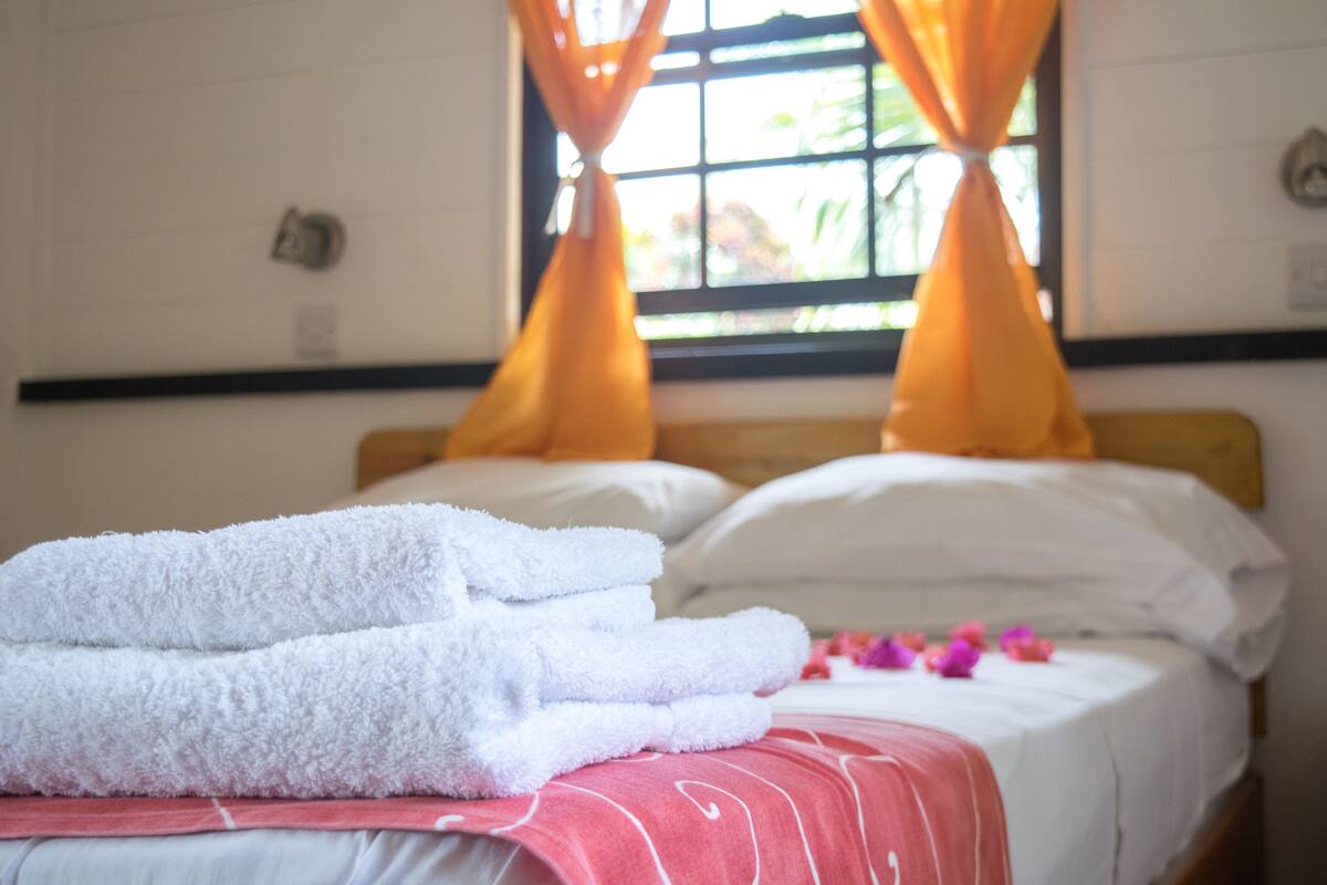 A neatly made bed with white pillows and a pink blanket, a pair of folded white towels, and a window with orange curtains.