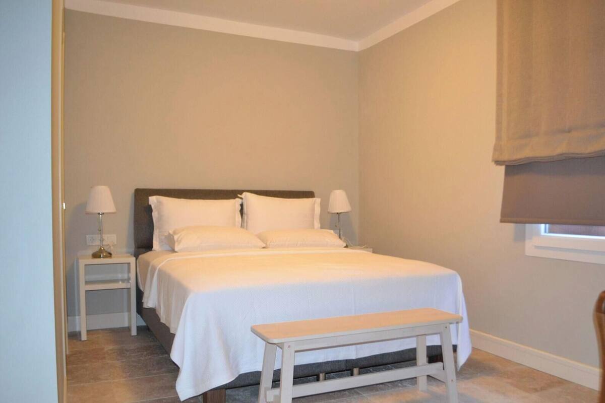 Ein Schlafzimmer mit einem Bett, Nachttischen, einer Bank und einem Fenster mit Jalousie.