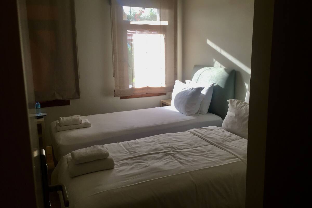 Ein Einzelbett mit weißer Bettwäsche, ein Nachttisch mit Lampe und ein Fenster mit Vorhängen.