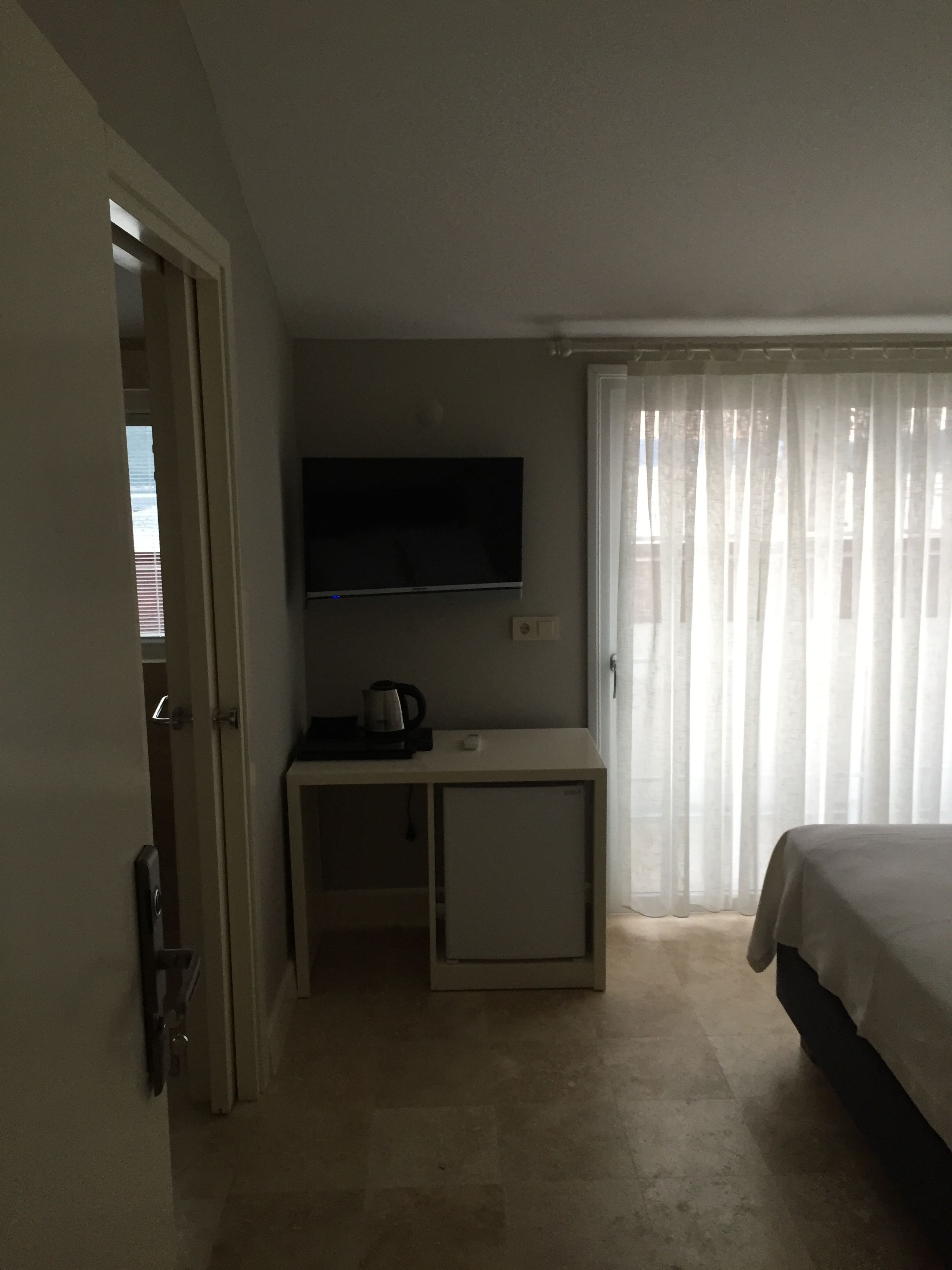 A hotel room with a bed, a TV mounted on the wall, a small desk with a kettle, and a door leading to another room.