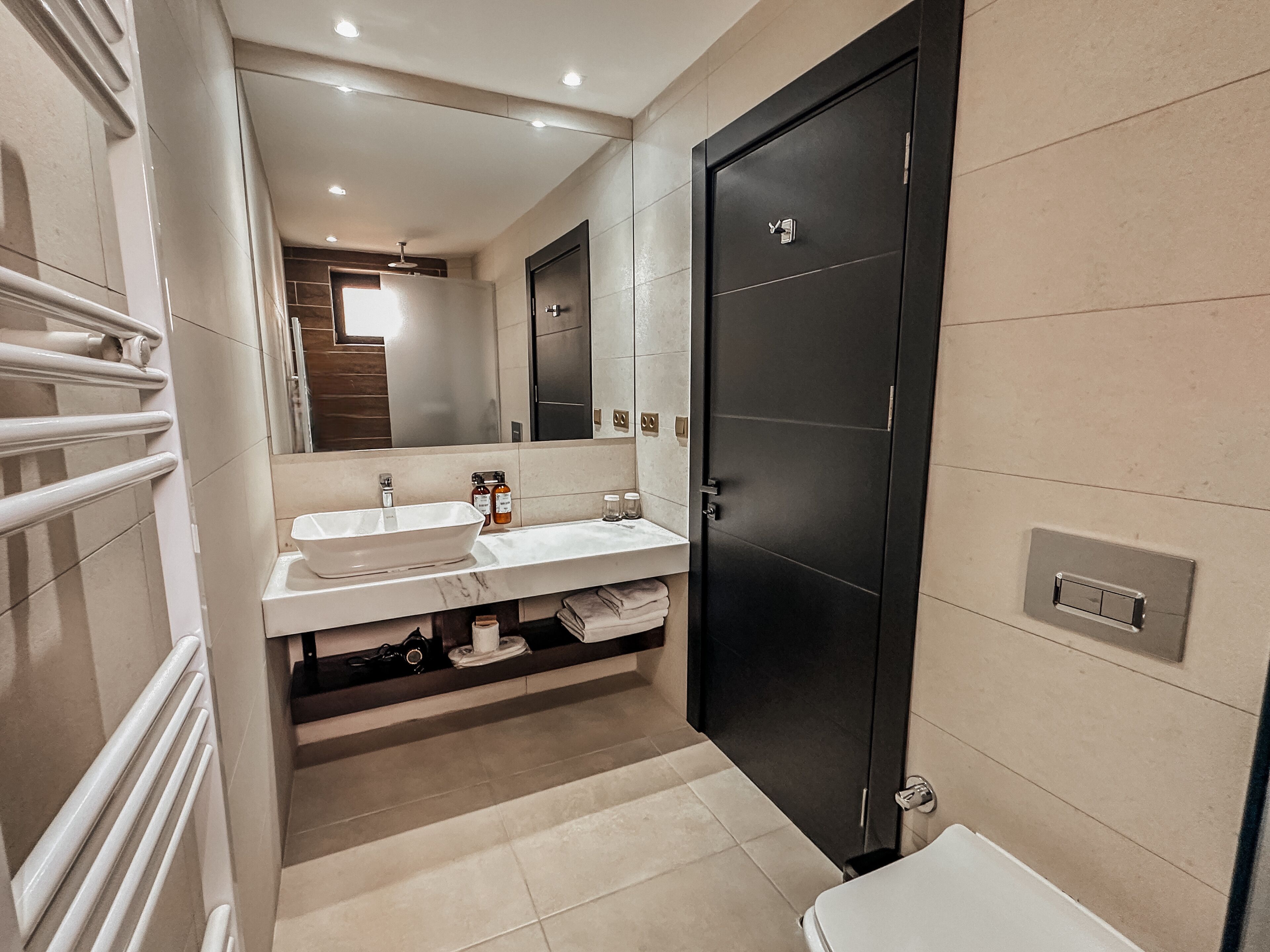 A modern bathroom with a large mirror, two sinks, a toilet, and a dark door.