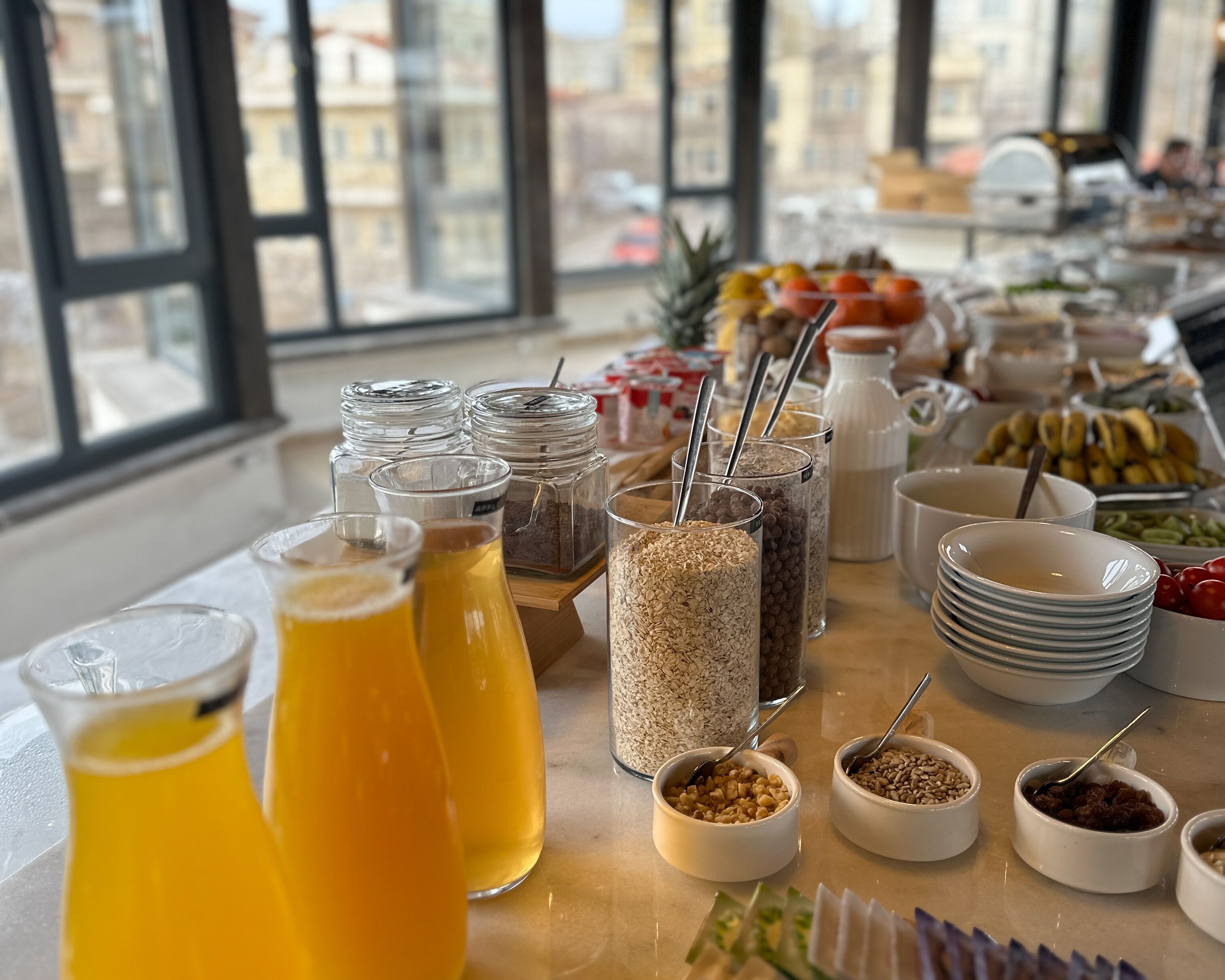 A breakfast buffet with various food items and beverages, including fruits, grains, and juices, arranged on a table with a city view in the background.