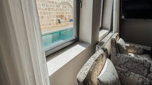 A room with a view of a swimming pool, stone wall, and a window with a curtain.