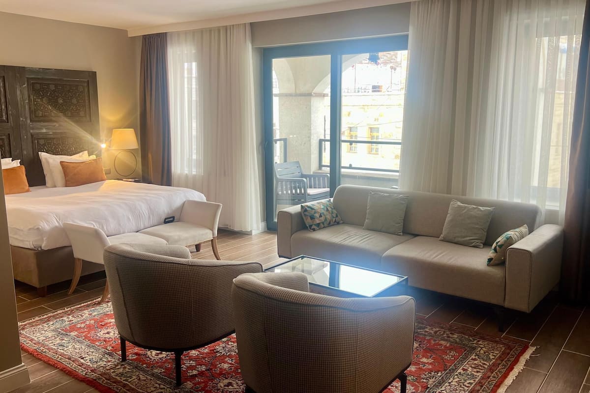 A hotel room with a bed, two armchairs, a sofa, a glass coffee table, and a view of the outside through large windows.