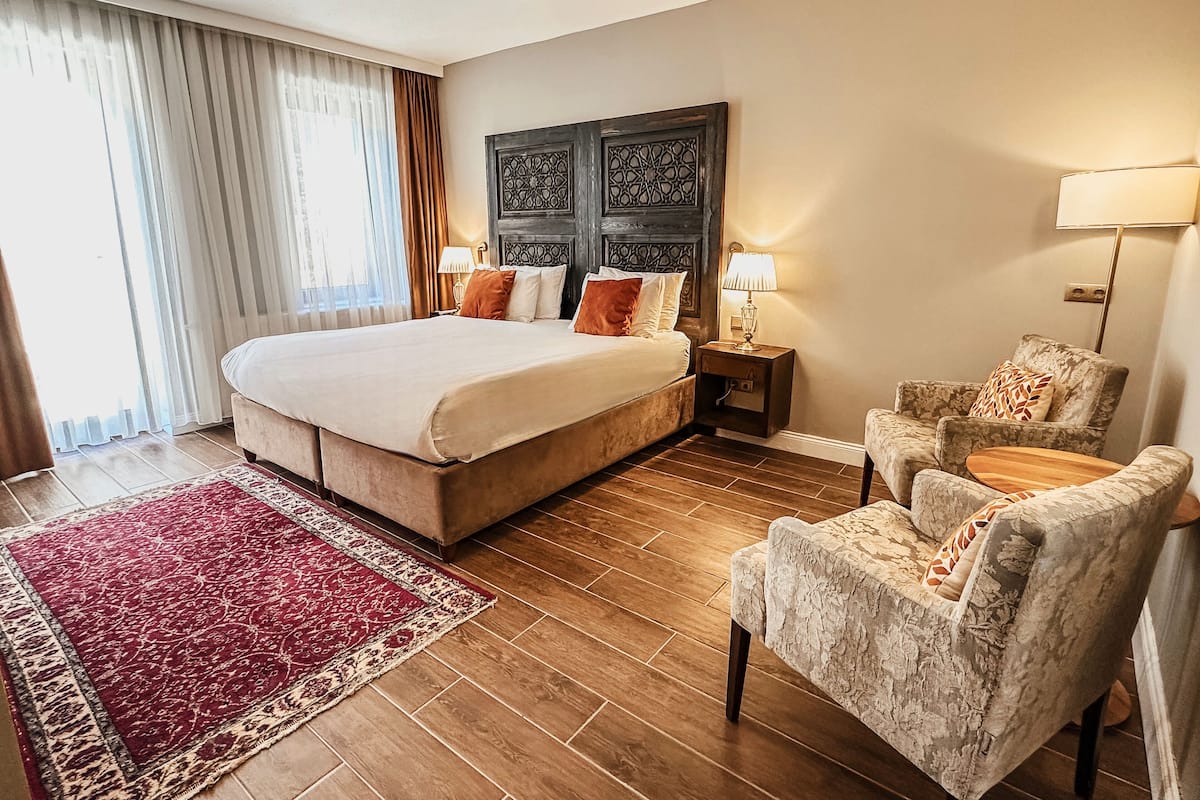 A hotel room with a large bed, two bedside tables with lamps, a floral armchair, a red and beige area rug, and a decorative headboard.