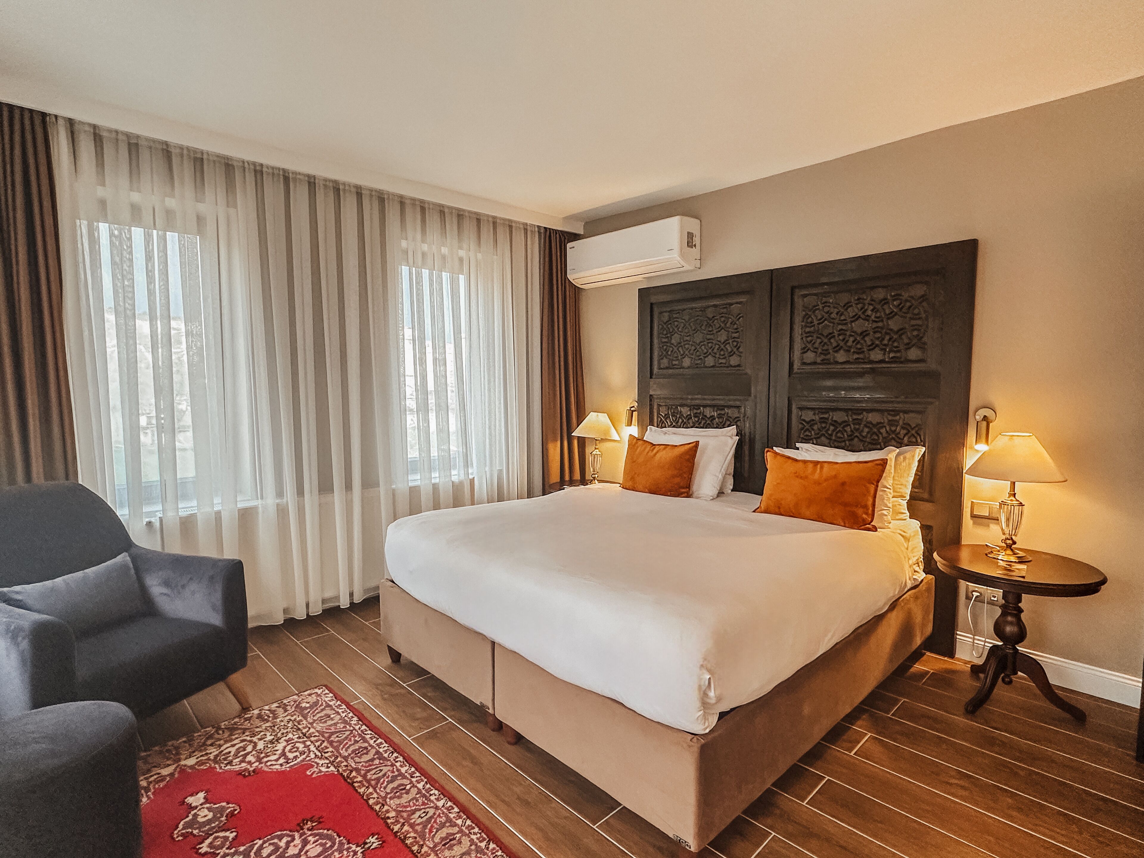 A hotel room with a large bed, two bedside lamps, a dark headboard, a red patterned rug, and a grey armchair.