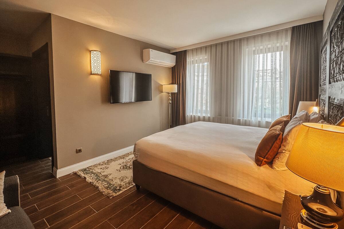 A hotel room with a large bed, a flat-screen TV mounted on the wall, a bedside table with a lamp, and a window with sheer curtains.